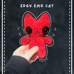 May include: A bright red crocheted cat plush toy with two large black button eyes and a stitched 'x' mouth. The text 'EDGY EMO CAT' is printed above the plush, which features four thin black whiskers extending from the sides of its face.