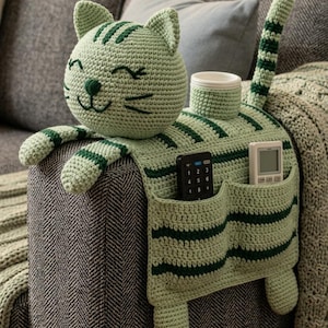 May include: A light green crochet cat-shaped sofa armrest organizer featuring dark green stripes, a head with embroidered facial features, a long tail, and two pockets containing a black remote control and a white digital remote control.