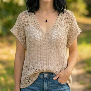 Può includere: Un top in maglia beige a maniche corte con motivo traforato, scollo a V e vestibilità rilassata. Il capo è abbinato a una semplice collana a catena dorata con un piccolo ciondolo scuro a forma di goccia che poggia sul petto.