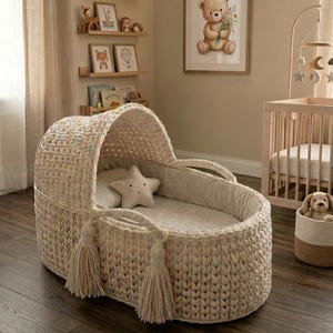May include: A cream-coloured, hand-crocheted baby bassinet featuring a rounded hood, two side handles with long decorative tassels, and a soft interior lining. Inside the bassinet rests a small, beige, star-shaped pillow on a flat mattress.