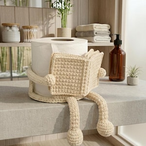May include: A beige crochet toilet paper holder featuring a figure with arms wrapped around a roll of toilet paper while holding an open book. The figure has two dangling legs with rounded feet, all crafted in a consistent light tan yarn texture.