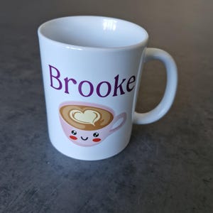 May include: White ceramic mug with the name "Brooke" in purple lettering. The mug features a cute cartoon coffee cup design with a heart-shaped foam design and smiling face. The mug has a white handle.