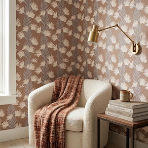 May include: A cream-coloured armchair with a textured fabric, draped with a rust-coloured woven throw blanket. Beside it, a stack of four books sits on a wooden side table, topped with a ceramic mug, against a brown wallpaper with a white leaf pattern.
