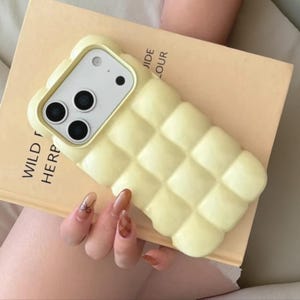 May include: A pale yellow phone case with a quilted design. The case has a camera cutout and is resting on a book with the text "WILD HERP" and "JIDE LOUR".