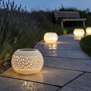 May include: Several white, spherical solar lanterns with leaf-shaped cutouts light up a stone path at dusk. The lanterns are spaced along the path, creating a warm glow. Lavender bushes and a wooden bench are visible in the background.