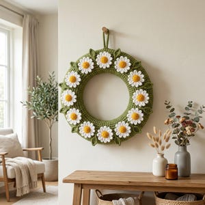 Puede incluir: Una corona circular de ganchillo con una base gruesa de color verde oliva adornada con once flores de margarita blancas con centros de color amarillo brillante. Las flores están espaciadas uniformemente alrededor del anillo.