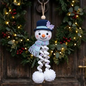 Op de afbeelding: Een gehaakte sneeuwpop-ornament met een donkerblauwe hoge hoed met een lichtblauwe band en een paarse bloem. De sneeuwpop heeft een oranje neus, een gestreepte sjaal en twee lange witte spiraalvormige benen die eindigen in pluizige witte pompons.
