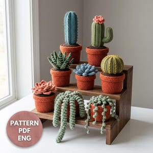 May include: A collection of eight crocheted plants in terracotta-colored pots, including a blue columnar cactus, a green cactus with a pink flower, a striped succulent, a blue rosette, a yellow cactus, and three trailing plants on a wooden stand.