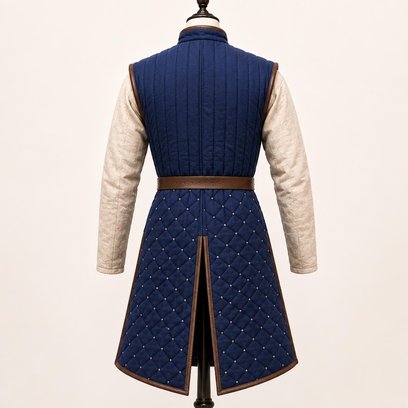May include: A long-sleeved blue quilted tunic featuring a vertical-stitched bodice and a diamond-patterned skirt section with small silver studs. The garment includes a brown leather belt at the waist and beige sleeves with a textured fabric finish.