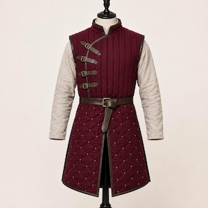 May include: A sleeveless, deep burgundy quilted vest featuring a high collar and a side-fastening design with four brown leather straps and brass buckles. The lower section has a diamond-stitched pattern with small studs and a brown leather belt.