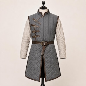 May include: A grey sleeveless tunic featuring vertical quilting on the chest and diamond-patterned quilting on the lower section. It includes four brown leather straps with brass buckles on the right side and a matching brown leather waist belt.