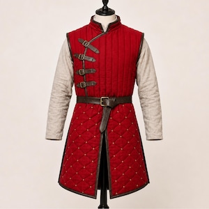 May include: A sleeveless red quilted vest featuring a high collar, vertical stitching on the chest, and a diamond-patterned skirt. The garment includes four dark brown leather straps with metal buckles on the side and a matching waist belt.