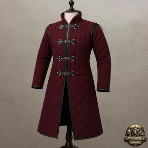 May include: A long-sleeved, knee-length quilted gambeson in a deep burgundy color. The garment features a high collar, black leather trim along the edges, and four vertical black leather straps with metal buckles down the center front closure.