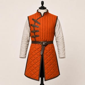 May include: A sleeveless orange quilted tunic featuring a high collar and a light-coloured long-sleeve shirt underneath. The tunic has a vertical row of four brown leather straps with metal buckles on the chest and a brown leather waist belt.