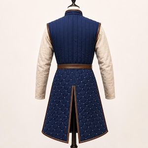 May include: A long-sleeved blue quilted tunic featuring a vertical-stitched bodice and a diamond-patterned skirt section with small silver studs. The garment includes a brown leather belt at the waist and beige sleeves with a textured fabric finish.