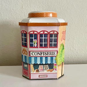 Peut inclure: Une bo&icirc;te d&eacute;corative en forme de p&acirc;tisserie fran&ccedil;aise. La bo&icirc;te pr&eacute;sente une fa&ccedil;ade rose avec des fen&ecirc;tres encadr&eacute;es de rouge, un auvent &agrave; rayures bleues et blanches et le mot "CONFISERIE". Le couvercle est blanc avec un dessus &agrave; motifs orange.