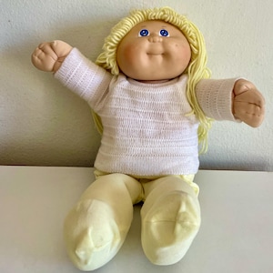 May include: A vintage Cabbage Patch Kid doll with blonde yarn hair and blue eyes. The doll is wearing a white sweater and yellow pants. The doll's arms are outstretched, and it is sitting down.