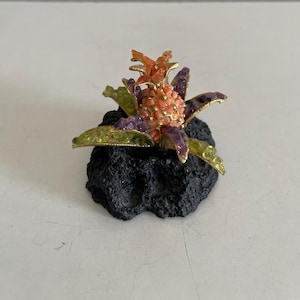 May include: A decorative sculpture featuring a central pineapple-shaped element with a textured orange surface, surrounded by pointed leaves in shades of purple and green, all mounted on a dark, porous, volcanic-looking black rock base.