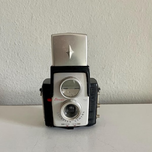 May include: A black and silver Kodak Brownie Starflex camera with a top-mounted silver flash unit. The front face features a white panel with the text Brownie, Dakon Lens, and Starflex Camera, along with settings for color and black and white film.
