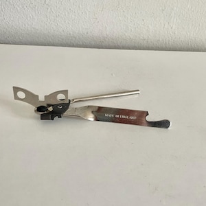 May include: A silver-colored can opener with a long handle and a cutting blade. The can opener has a black gear mechanism and two small holes on the top. The text "MADE IN ENGLAND" is printed on the blade.
