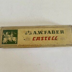 May include: A rectangular, vintage metal pencil tin with a cream-colored finish. The lid features a green square with a knight on horseback, the text A.W. FABER CASTELL in black and red, a scale icon, and the text A.W. FABER CASTELL STEIN BEI N&Uuml;RNBERG.