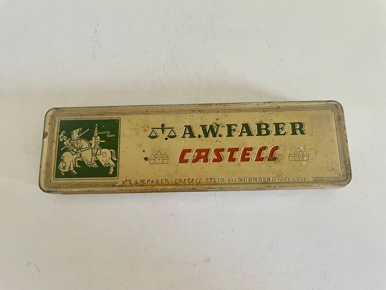 May include: A rectangular, vintage metal pencil tin with a cream-colored finish. The lid features a green square with a knight on horseback, the text A.W. FABER CASTELL in black and red, a scale icon, and the text A.W. FABER CASTELL STEIN BEI N&Uuml;RNBERG.