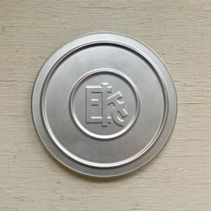 May include: A round, silver-colored metal lid with a raised edge. The center features a circular design with Japanese characters. The lid is likely for a container, possibly for food or storage.