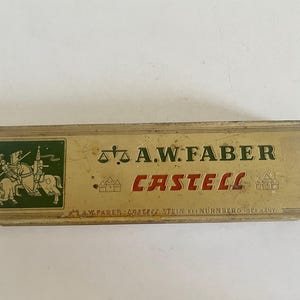 May include: A rectangular, vintage metal pencil tin with a gold-toned finish. The lid features a green square with a knight on horseback, a scale icon, and the text A.W. FABER CASTELL. Small castle icons flank the red text, with A.W. FABER CASTELL STEIN BEI N&Uuml;RNBERG (GERMANY) printed at the bottom.