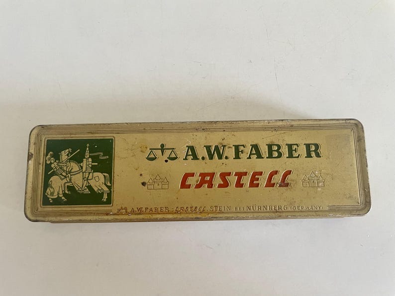 May include: A rectangular, vintage metal pencil tin with a gold-toned finish. The lid features a green square with a knight on horseback, a scale icon, and the text A.W. FABER CASTELL. Small castle icons flank the red text, with A.W. FABER CASTELL STEIN BEI N&Uuml;RNBERG (GERMANY) printed at the bottom.