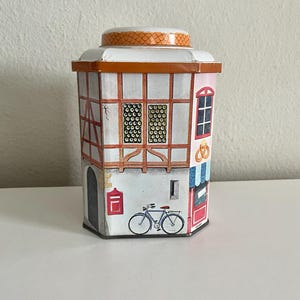 Peut inclure: Une bo&icirc;te d&eacute;corative en forme de b&acirc;timent europ&eacute;en. La bo&icirc;te pr&eacute;sente une fa&ccedil;ade blanche avec un encadrement en bois brun, une bo&icirc;te aux lettres rouge et un v&eacute;lo. Le toit est orange avec un motif textur&eacute;. Des fen&ecirc;tres et une porte sont &eacute;galement visibles.