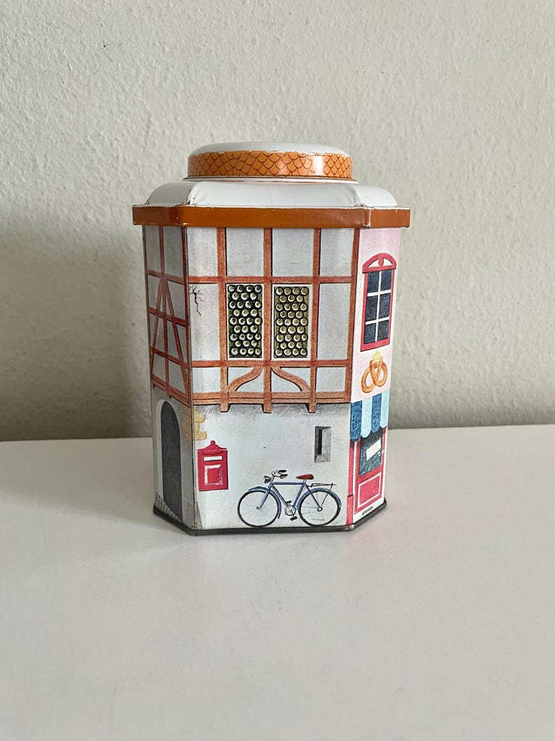 Peut inclure: Une bo&icirc;te d&eacute;corative en forme de b&acirc;timent europ&eacute;en. La bo&icirc;te pr&eacute;sente une fa&ccedil;ade blanche avec un encadrement en bois brun, une bo&icirc;te aux lettres rouge et un v&eacute;lo. Le toit est orange avec un motif textur&eacute;. Des fen&ecirc;tres et une porte sont &eacute;galement visibles.