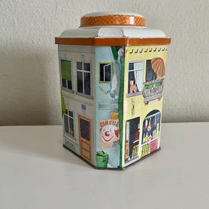 Peut inclure: Bo&icirc;te en &eacute;tain vintage en forme de b&acirc;timent, avec des illustrations color&eacute;es de fen&ecirc;tres, de balcons et d'une affiche de cirque. Le couvercle est blanc avec une bordure orange &agrave; motifs. Forme rectangulaire.