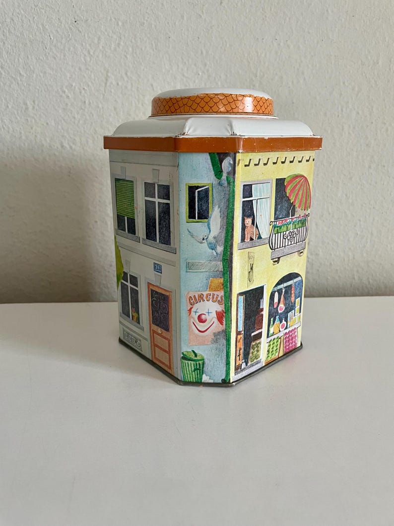 Peut inclure: Bo&icirc;te en &eacute;tain vintage en forme de b&acirc;timent, avec des illustrations color&eacute;es de fen&ecirc;tres, de balcons et d'une affiche de cirque. Le couvercle est blanc avec une bordure orange &agrave; motifs. Forme rectangulaire.