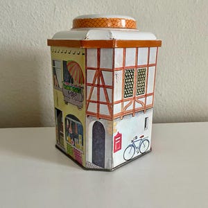 Peut inclure: Une bo&icirc;te en m&eacute;tal vintage en forme de b&acirc;timent europ&eacute;en. La bo&icirc;te pr&eacute;sente des illustrations d'un balcon avec un parapluie, une porte, des fen&ecirc;tres et un v&eacute;lo. Le toit est blanc avec une bordure orange.