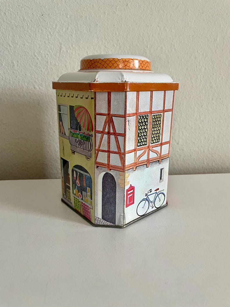 Peut inclure: Une bo&icirc;te en m&eacute;tal vintage en forme de b&acirc;timent europ&eacute;en. La bo&icirc;te pr&eacute;sente des illustrations d'un balcon avec un parapluie, une porte, des fen&ecirc;tres et un v&eacute;lo. Le toit est blanc avec une bordure orange.