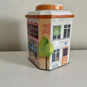 Peut inclure: Une bo&icirc;te en m&eacute;tal d&eacute;corative en forme de b&acirc;timent, repr&eacute;sentant une fa&ccedil;ade de confiserie avec le mot "CONFISERIE". La bo&icirc;te est blanche avec des bordures orange, un couvercle bomb&eacute; et un arbre peint. Le b&acirc;timent a des fen&ecirc;tres et une porte.