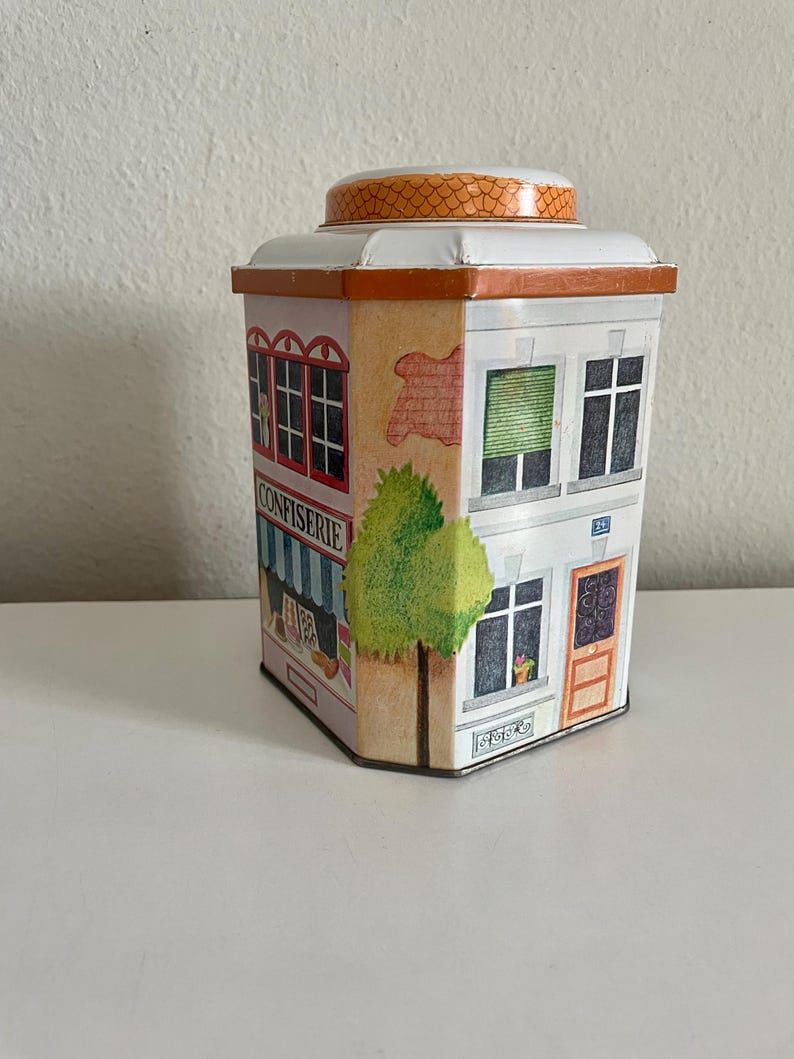 Peut inclure: Une bo&icirc;te en m&eacute;tal d&eacute;corative en forme de b&acirc;timent, repr&eacute;sentant une fa&ccedil;ade de confiserie avec le mot "CONFISERIE". La bo&icirc;te est blanche avec des bordures orange, un couvercle bomb&eacute; et un arbre peint. Le b&acirc;timent a des fen&ecirc;tres et une porte.