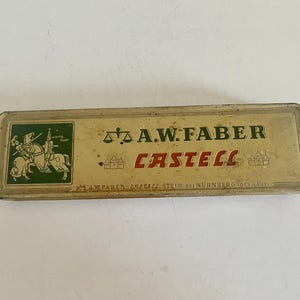 May include: A rectangular, vintage metal pencil case in a muted gold tone. The lid features a green square with a knight on horseback, a scale icon, and the text A.W. FABER CASTELL. Small castle icons flank the red text, with fine print at the bottom.