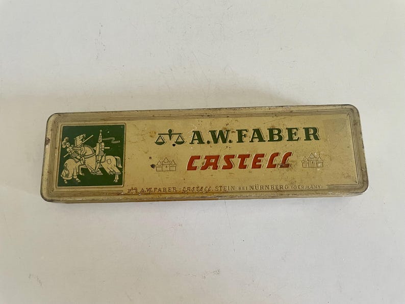 May include: A rectangular, vintage metal pencil case in a muted gold tone. The lid features a green square with a knight on horseback, a scale icon, and the text A.W. FABER CASTELL. Small castle icons flank the red text, with fine print at the bottom.