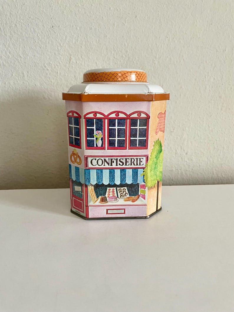 Peut inclure: Une bo&icirc;te d&eacute;corative en forme de p&acirc;tisserie fran&ccedil;aise. La bo&icirc;te pr&eacute;sente une fa&ccedil;ade rose avec des fen&ecirc;tres encadr&eacute;es de rouge, un auvent &agrave; rayures bleues et blanches et le mot "CONFISERIE". Le couvercle est blanc avec un dessus &agrave; motifs orange.