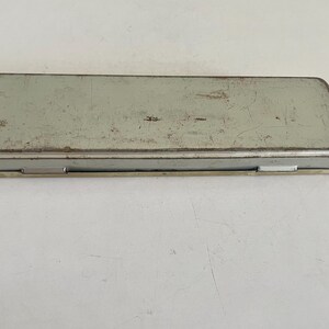 May include: A rectangular, light gray metal pencil case with a hinged lid. The surface of the container shows visible signs of wear, including scratches, scuffs, and minor rust along the edges, indicating a weathered and aged appearance.