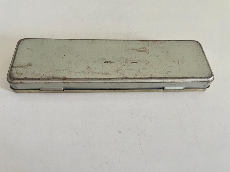 May include: A rectangular, light gray metal pencil case with a hinged lid. The surface of the container shows visible signs of wear, including scratches, scuffs, and minor rust along the edges, indicating a weathered and aged appearance.