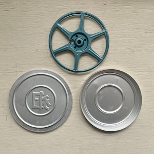 May include: An overhead shot features a teal-colored film reel and two silver metal film canister lids. One lid has an embossed Japanese character. The items are arranged on a light-colored wooden surface.