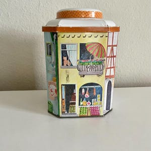 Peut inclure: Une bo&icirc;te en &eacute;tain vintage avec un design fantaisiste repr&eacute;sentant une sc&egrave;ne de village europ&eacute;en. Le r&eacute;cipient pr&eacute;sente un cirque, une boutique avec des produits et un balcon avec un chat. Le couvercle est orange et le r&eacute;cipient est &agrave; plusieurs faces.