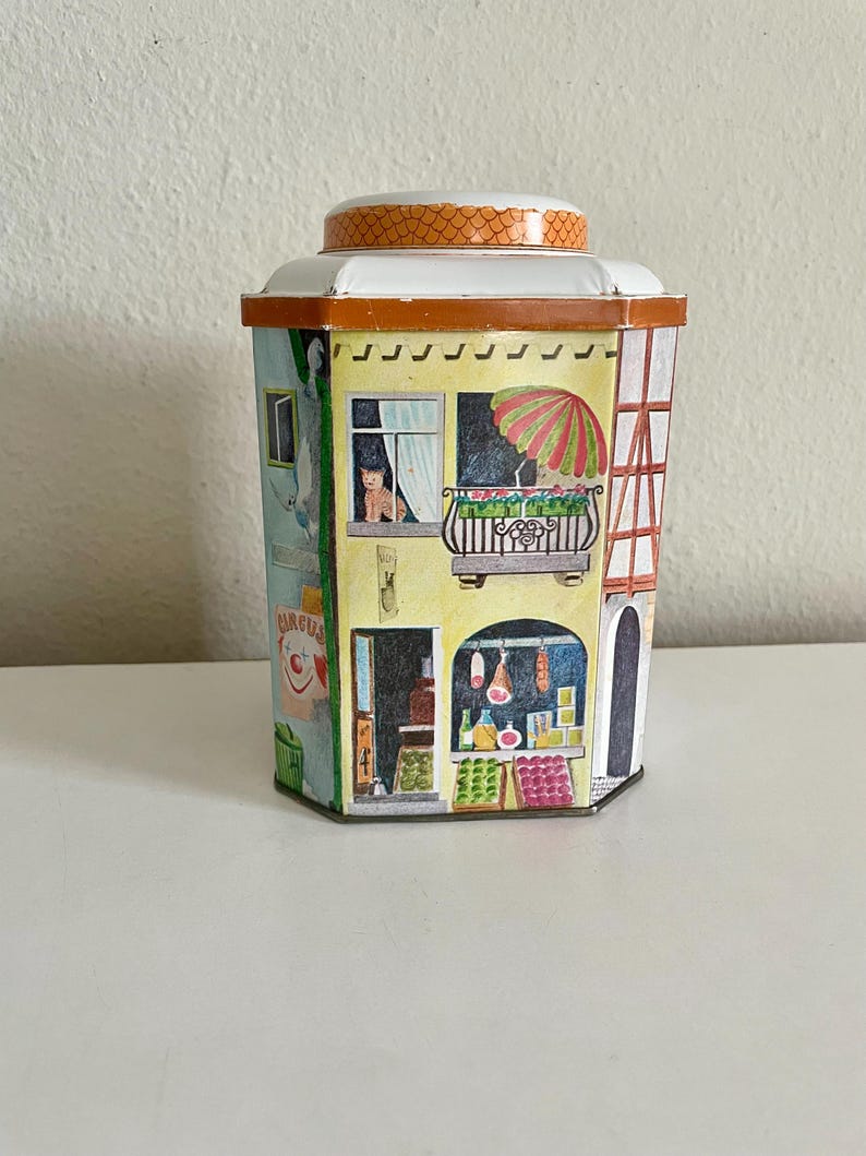 Peut inclure: Une bo&icirc;te en &eacute;tain vintage avec un design fantaisiste repr&eacute;sentant une sc&egrave;ne de village europ&eacute;en. Le r&eacute;cipient pr&eacute;sente un cirque, une boutique avec des produits et un balcon avec un chat. Le couvercle est orange et le r&eacute;cipient est &agrave; plusieurs faces.