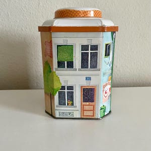Peut inclure: Bo&icirc;te en m&eacute;tal d&eacute;corative en forme de b&acirc;timent, avec des illustrations de fen&ecirc;tres, de portes et d'un arbre. La bo&icirc;te a un couvercle blanc avec une bordure orange et un dessus &agrave; motifs. La bo&icirc;te a une vari&eacute;t&eacute; de couleurs, dont l'orange, le vert, le bleu et le marron.