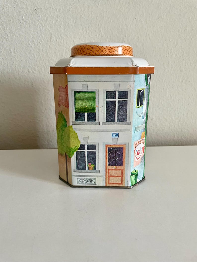 Peut inclure: Bo&icirc;te en m&eacute;tal d&eacute;corative en forme de b&acirc;timent, avec des illustrations de fen&ecirc;tres, de portes et d'un arbre. La bo&icirc;te a un couvercle blanc avec une bordure orange et un dessus &agrave; motifs. La bo&icirc;te a une vari&eacute;t&eacute; de couleurs, dont l'orange, le vert, le bleu et le marron.