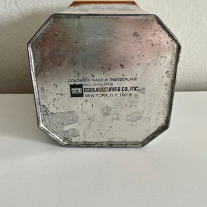 Peut inclure: Un r&eacute;cipient m&eacute;tallique vintage avec une base octogonale. Le r&eacute;cipient a une finition argent&eacute;e avec des signes d'usure. Le texte "CONTAINER MADE IN SWITZERLAND EXCLUSIVELY FOR CAME MANUFACTURING CO., INC NEW YORK, N.Y. 10013" est imprim&eacute; sur la base.