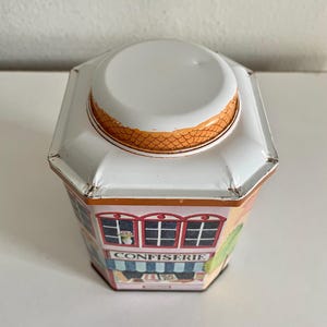 Peut inclure: Bo&icirc;te en m&eacute;tal octogonale avec couvercle blanc et illustration color&eacute;e d'une boutique. Le mot "CONFISERIE" est inscrit sur le devant. Le bord du couvercle pr&eacute;sente un motif en &eacute;cailles orange.