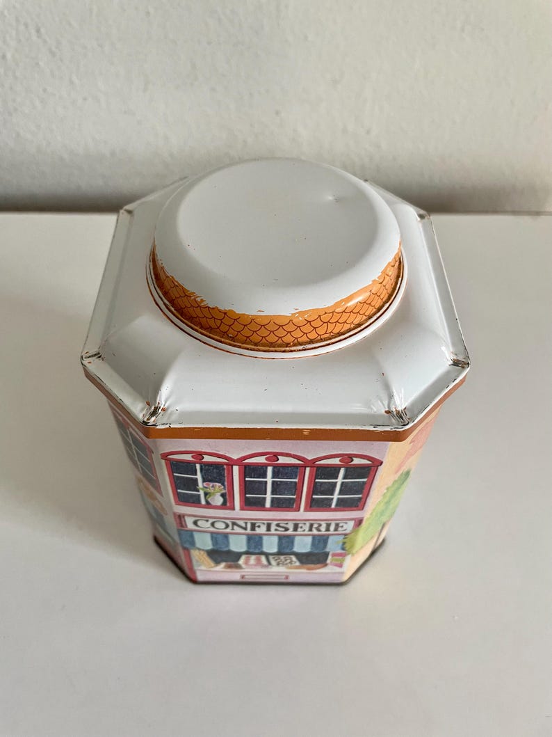 Peut inclure: Bo&icirc;te en m&eacute;tal octogonale avec couvercle blanc et illustration color&eacute;e d'une boutique. Le mot "CONFISERIE" est inscrit sur le devant. Le bord du couvercle pr&eacute;sente un motif en &eacute;cailles orange.